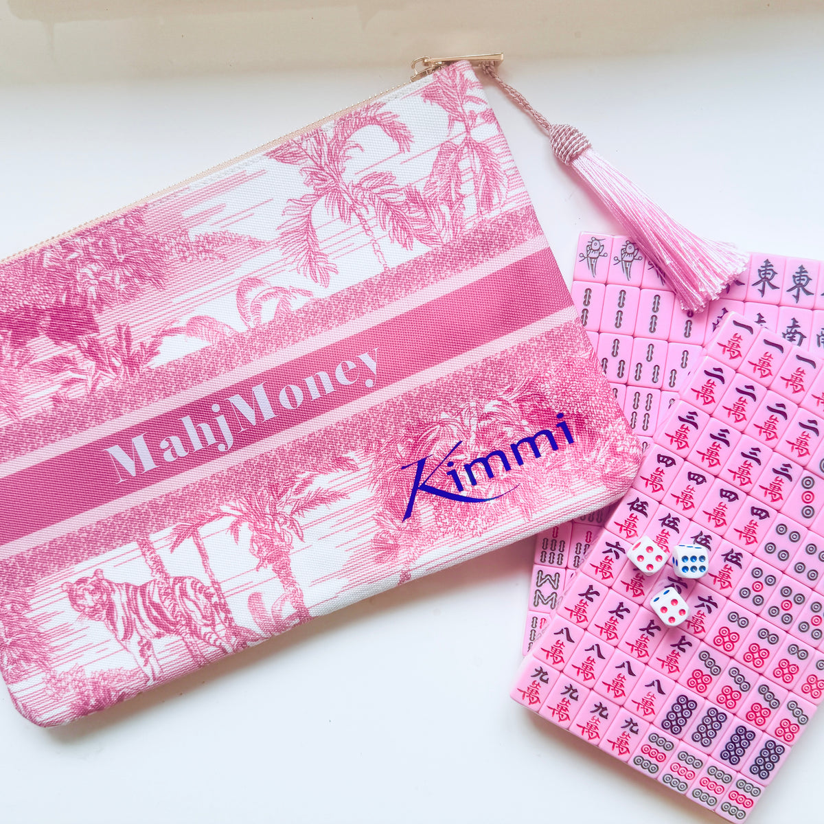 A pink and white tropical-themed zippered pouch with a tassel, featuring the text 'MahjMoney' and 'Kimmi', displayed next to a pink mahjong tile card deck and two dice on a white surface.